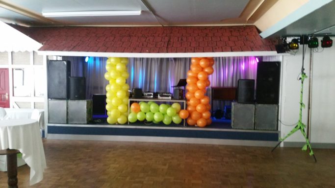 Bunte Ballondekoration in Gelb, Orange und Grün an einer Bühne mit Lautsprechern.